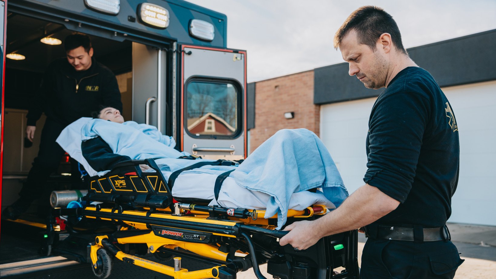 Two paramedics transporting a patient on a stretcher into an ambulance outside a building.