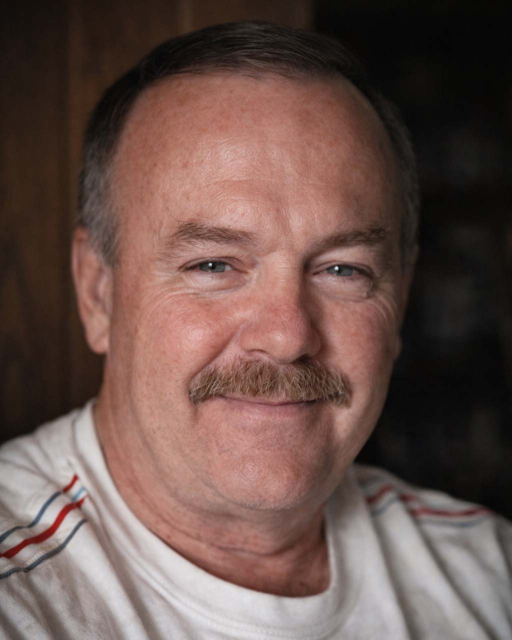 Close-up portrait of a middle-aged man with a mustache smiling, wearing a white shirt with red and blue stripes on the shoulder.