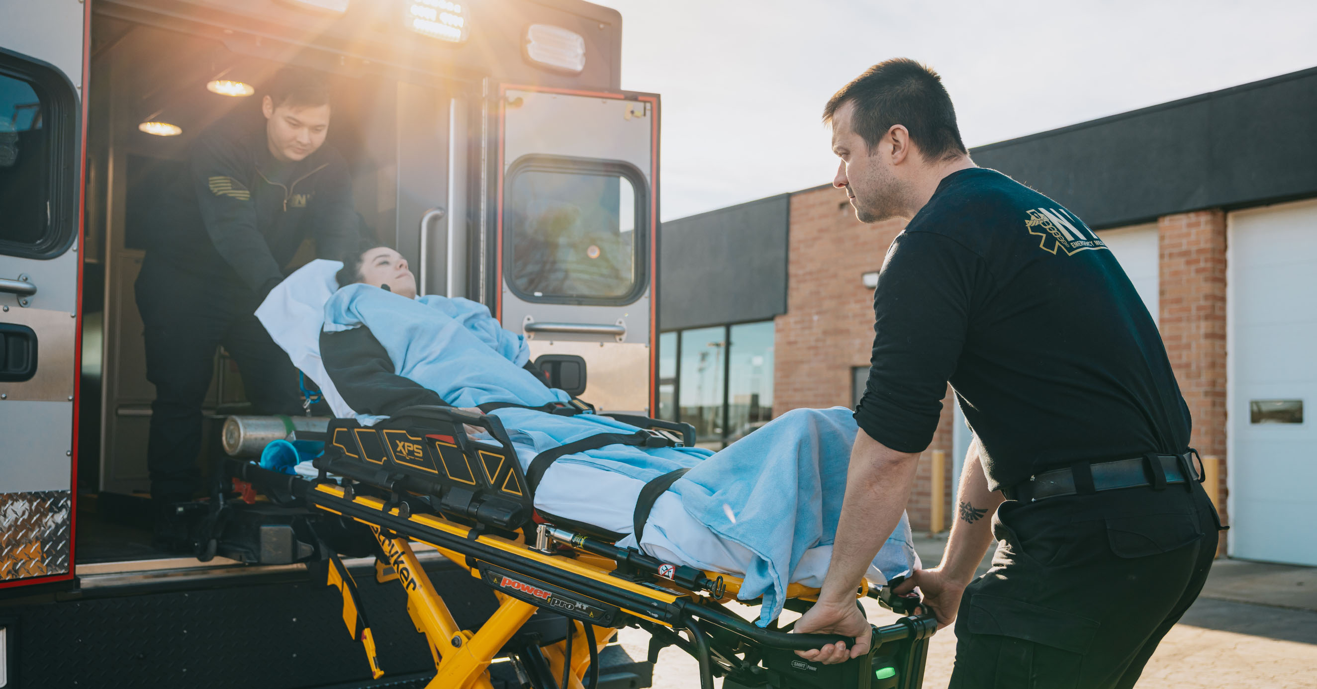 Two emergency medical technicians loading a patient covered with a blue blanket into an ambulance on a yellow stretcher.