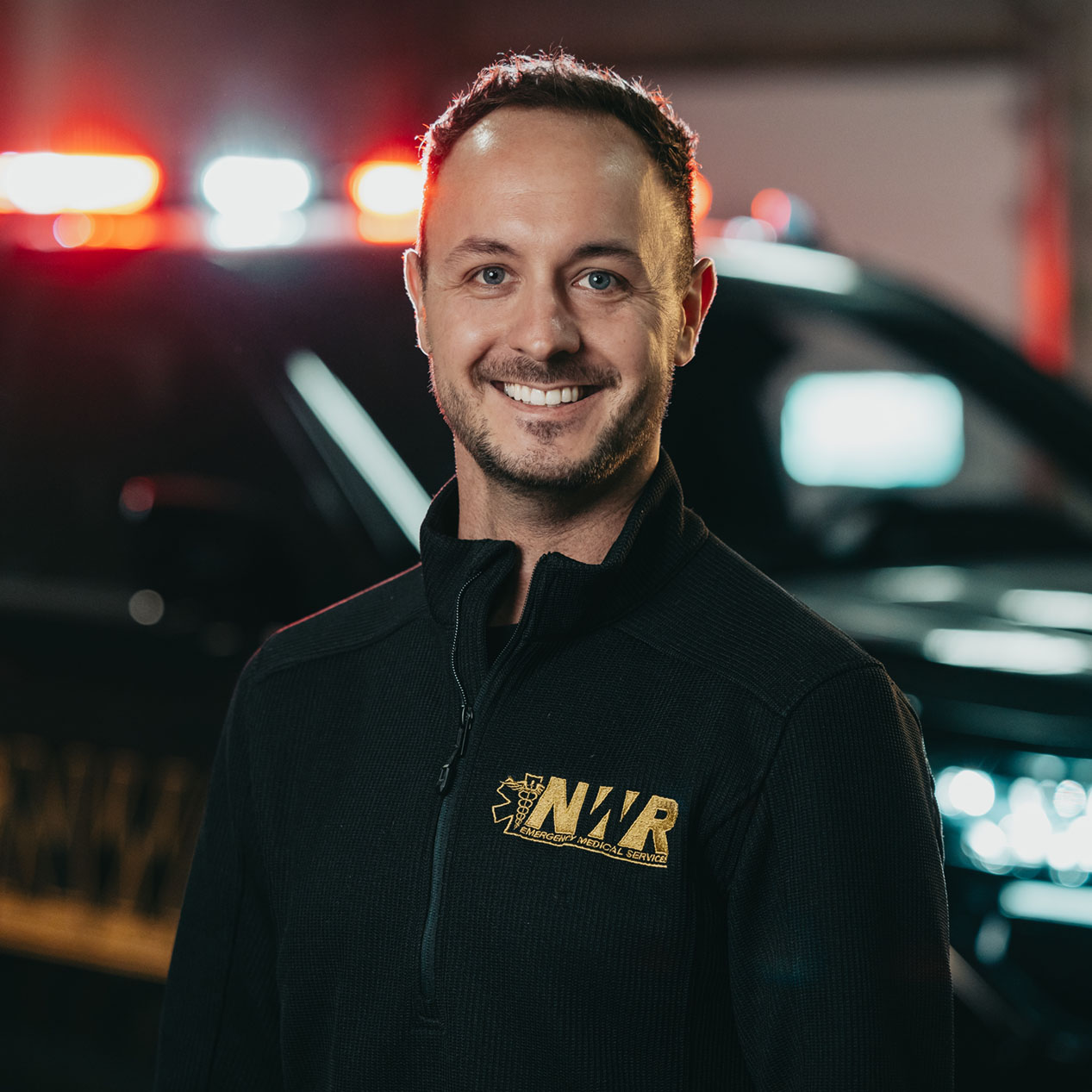Smiling man wearing a black NW Rescue EMS jacket standing in front of a vehicle with emergency lights.