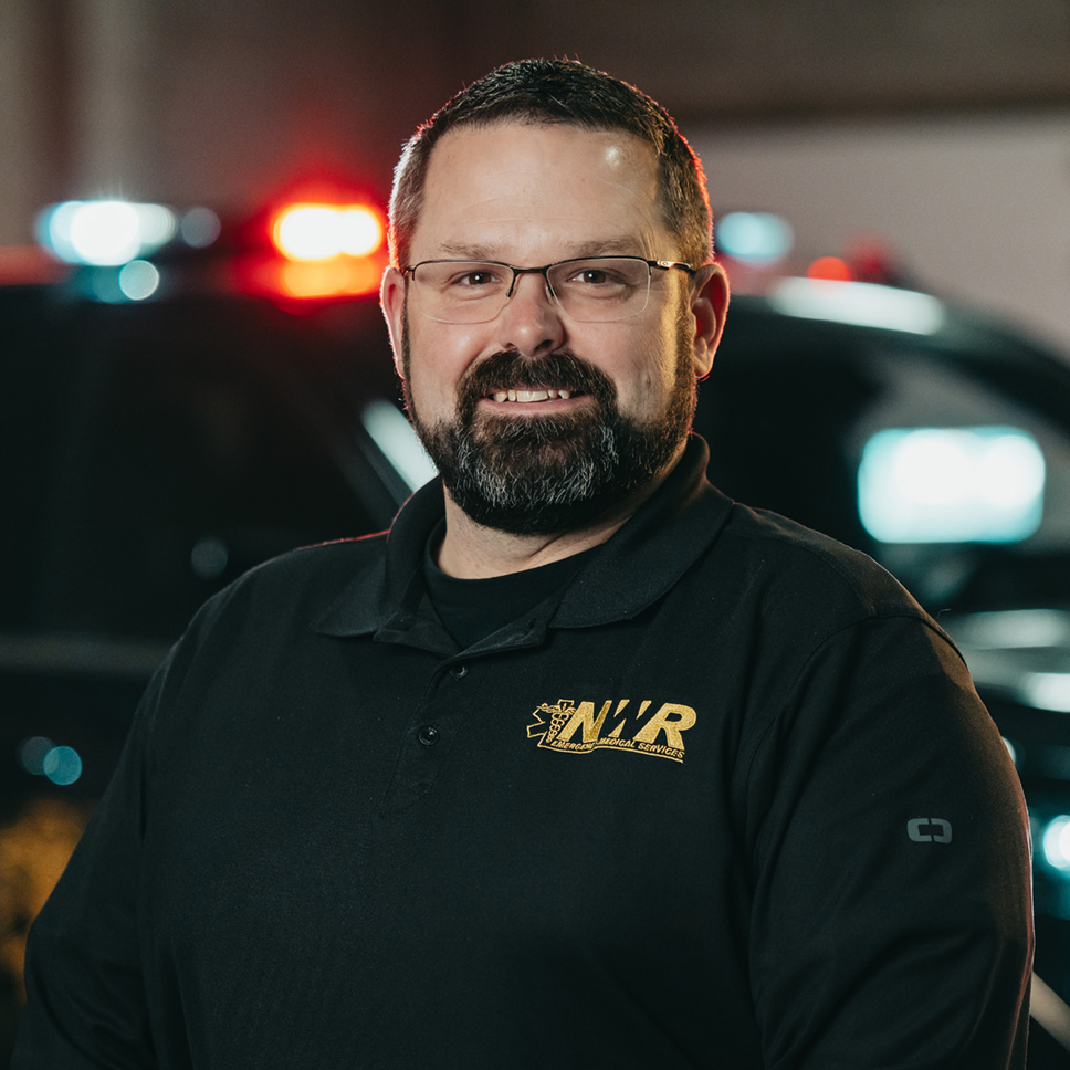 Smiling man with glasses and beard wearing a black NWR Emergency Medical Services shirt in front of a police vehicle with red and blue lights.