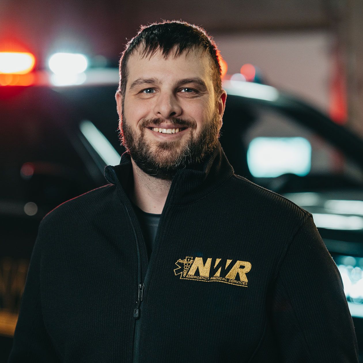 Smiling man with beard wearing a black jacket with NWR Emergency Medical Services logo, standing in front of a vehicle with flashing red and blue lights.