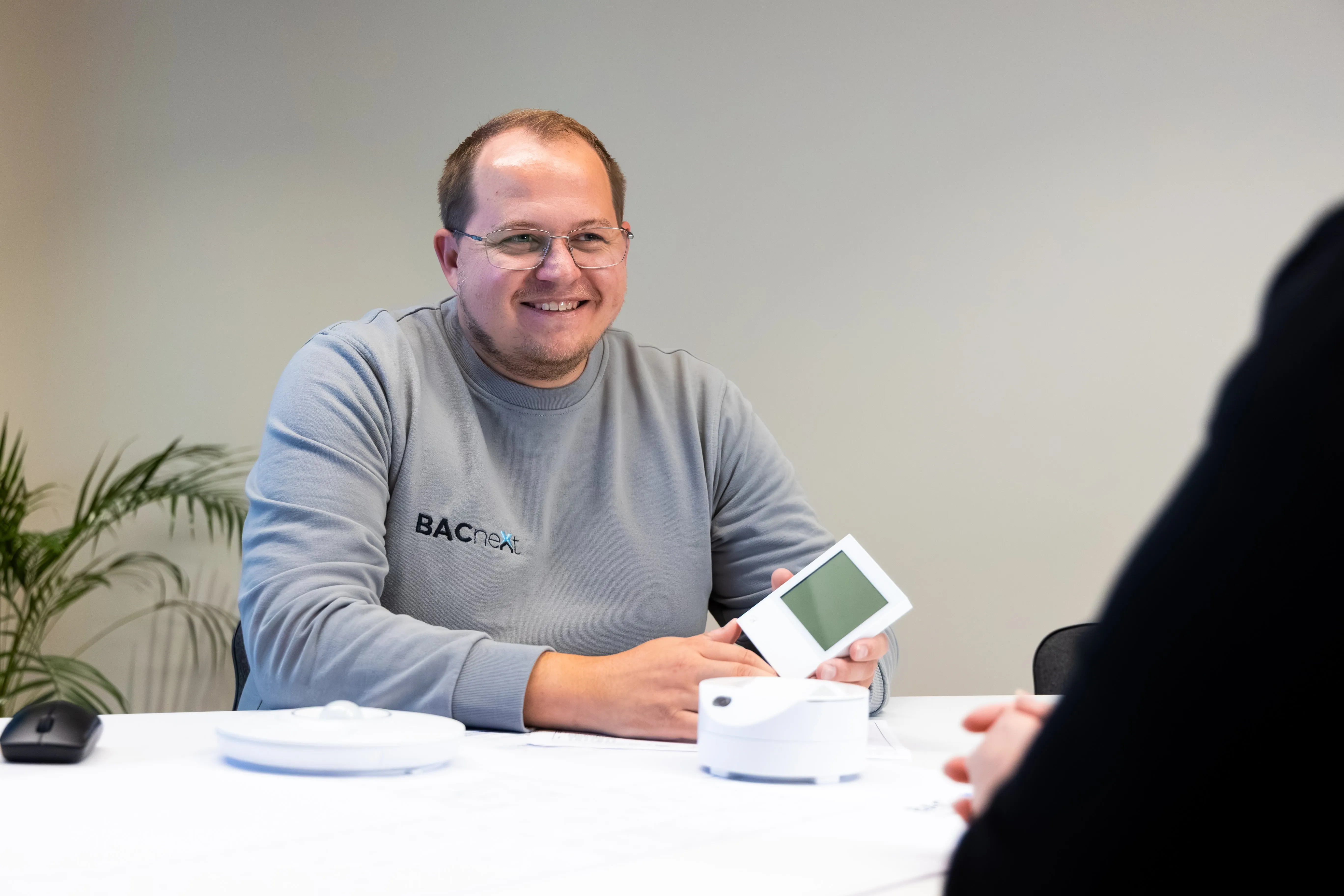 Ein lächelnder Mann mit grauem BACnet-Pullover zeigt ein elektronisches Gerät mit Bildschirm an einem weißen Tisch.