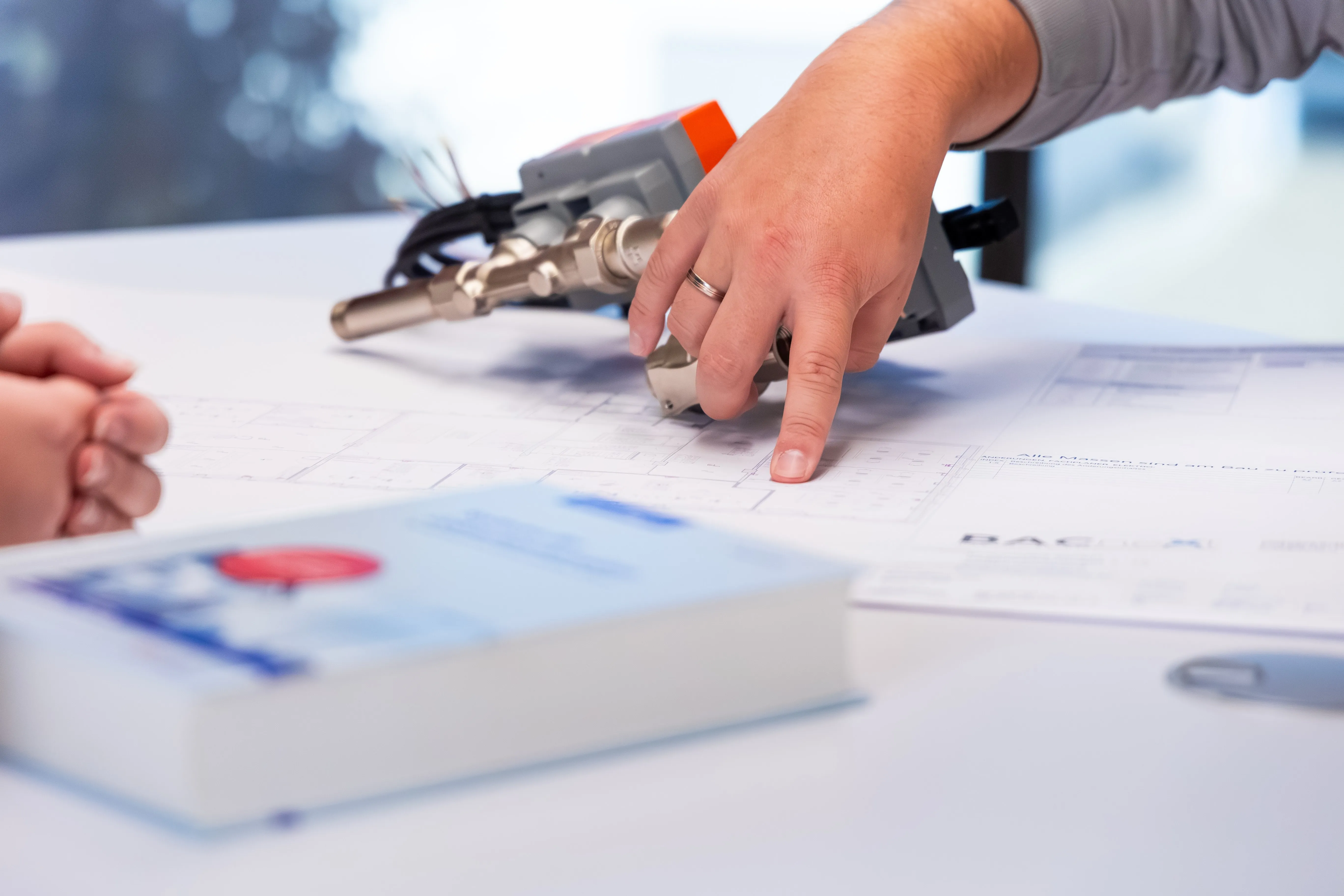 Eine Hand zeigt auf eine technische Zeichnung auf einem Tisch; im Vordergrund sind ein mechanisches Gerät und ein verschwommenes Buch zu sehen.