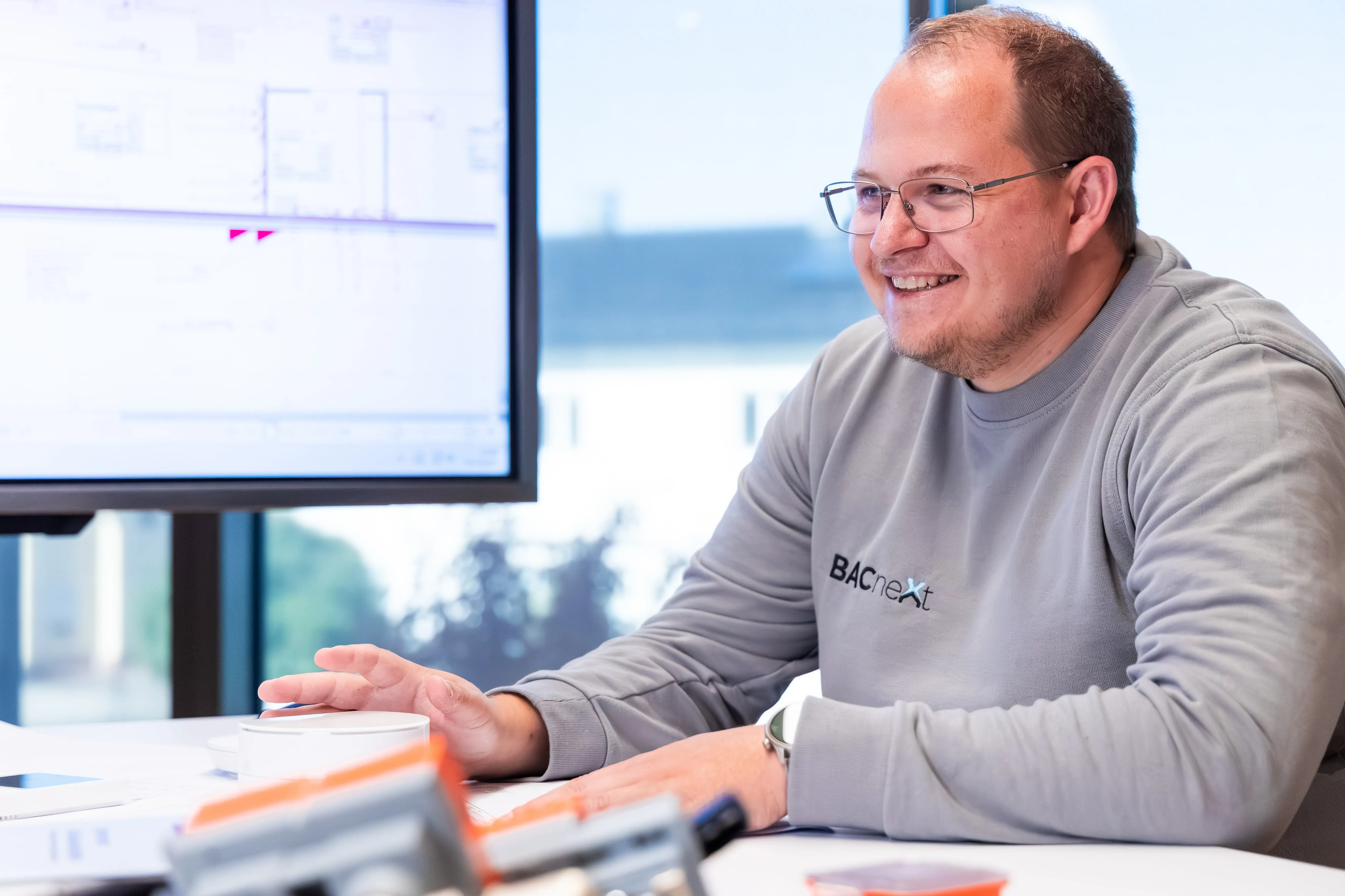 Mann mit Brille und grauem BACnet-Sweatshirt sitzt lächelnd an einem Tisch mit technischen Geräten und Dokumenten.