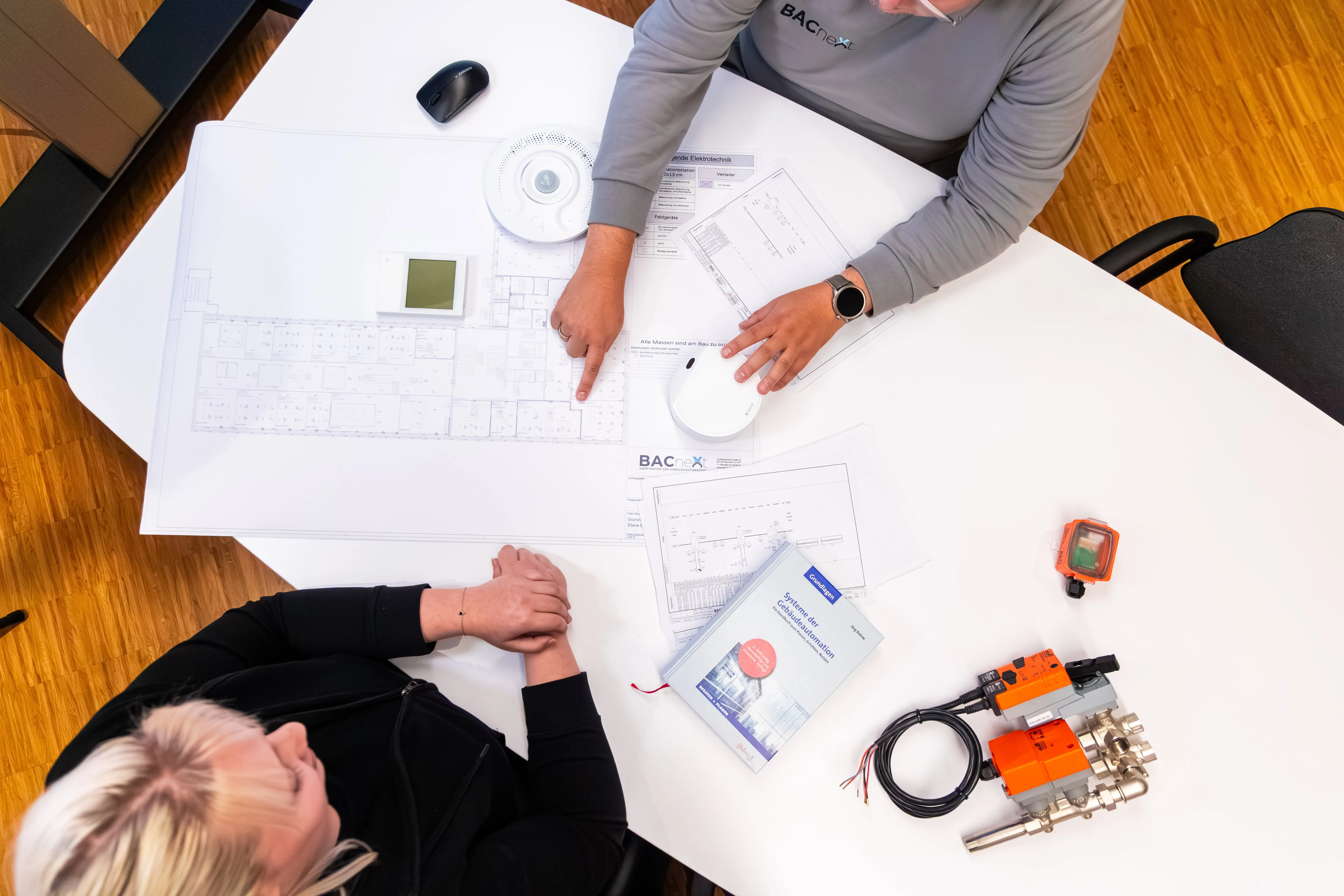 Zwei Personen besprechen Baupläne und technische Gebäudeautomations-Planungen auf einem weißen Tisch mit Holzboden.