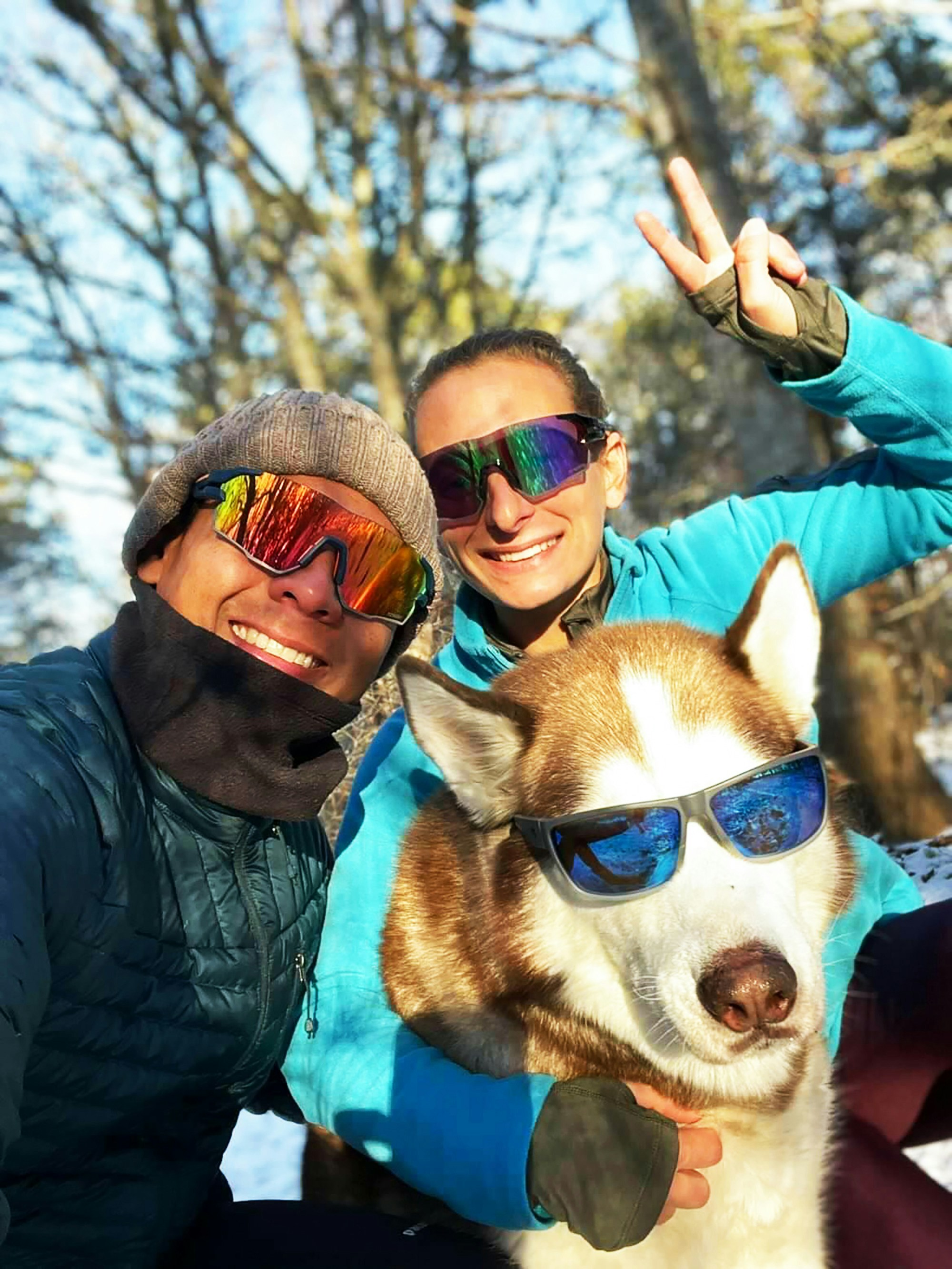 Photographie amusante de deux personnes et un chien de race Husky, tous les trois portant des lunettes de soleil, posant à l'extérieur.