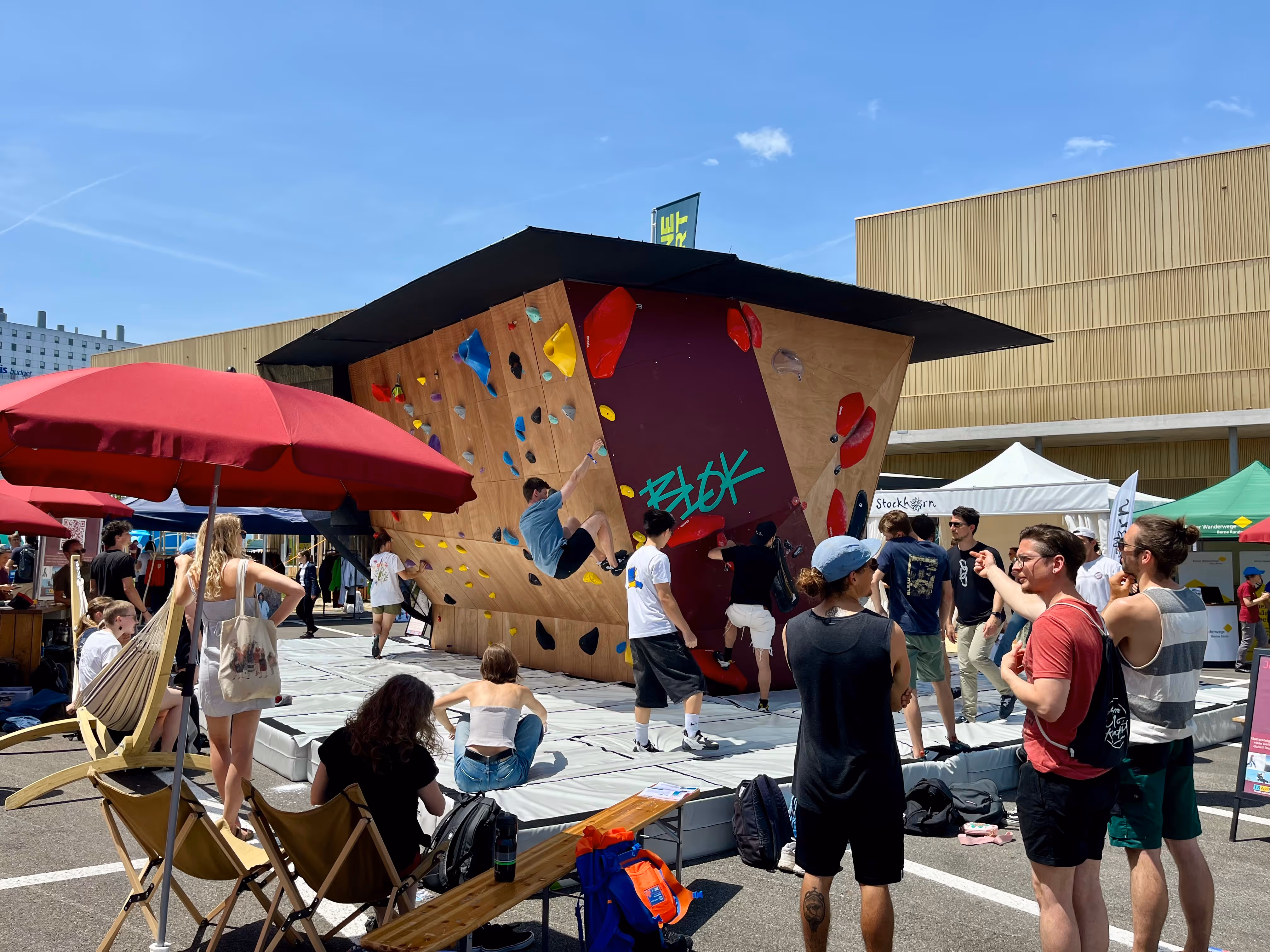 Outdoor climbing event with people watching and participating on a bouldering wall under a black canopy during a sunny day.