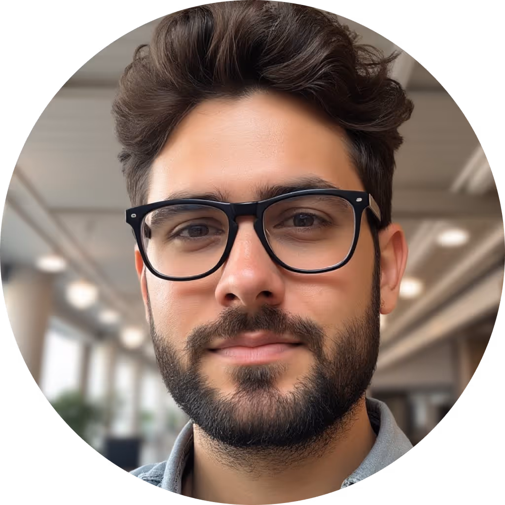 Portrait of a young man with dark hair, beard, and black rectangular glasses in an indoor setting.