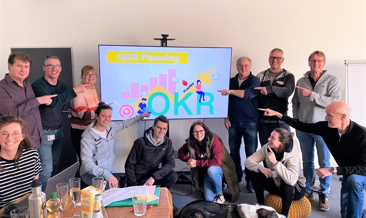 Teilnehmende eines OKR-Workshops stehen und sitzen gemeinsam vor einem Bildschirm mit der Aufschrift „OKR Planning“ und zeigen auf die dargestellte OKR-Grafik. Die Gruppe wirkt engagiert und motiviert und symbolisiert die gemeinsame Arbeit an Zielen, Prioritäten und strategischer Umsetzung.