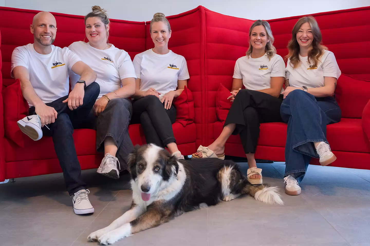 Fünf Personen in weißen T-Shirts sitzen auf rotem Sofa, ein Hund liegt vorne auf dem Boden.
