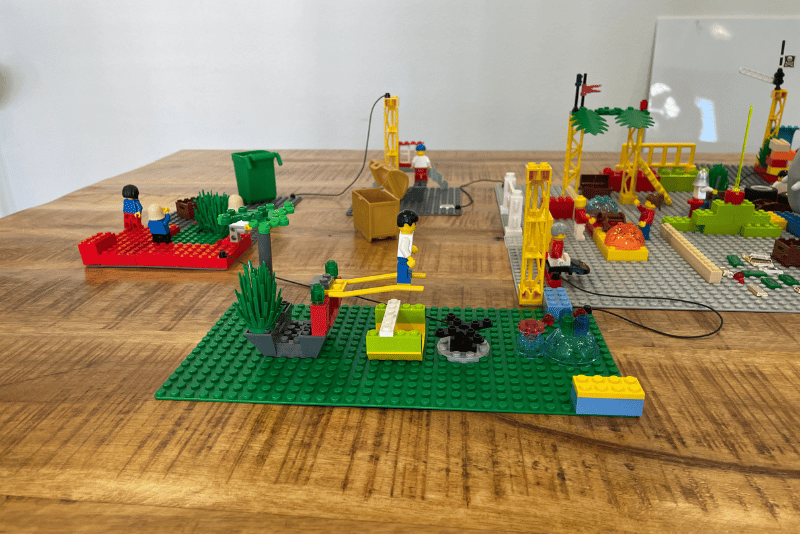 Fünf Personen bauen mit Legosteinen an einem Tisch in einem hellen Raum mit Fenster und Leinwand.