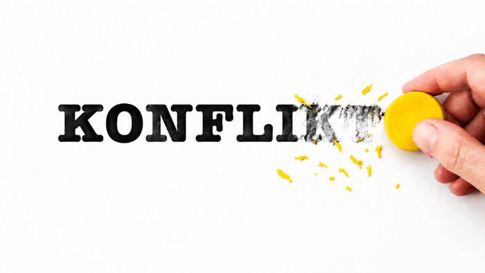 Eine Hand radiert mit einem runden, gelben Radiergummi das Wort „KONFLIKT“ auf weißem Hintergrund teilweise aus – als Symbol für die Lösung von Konflikten.