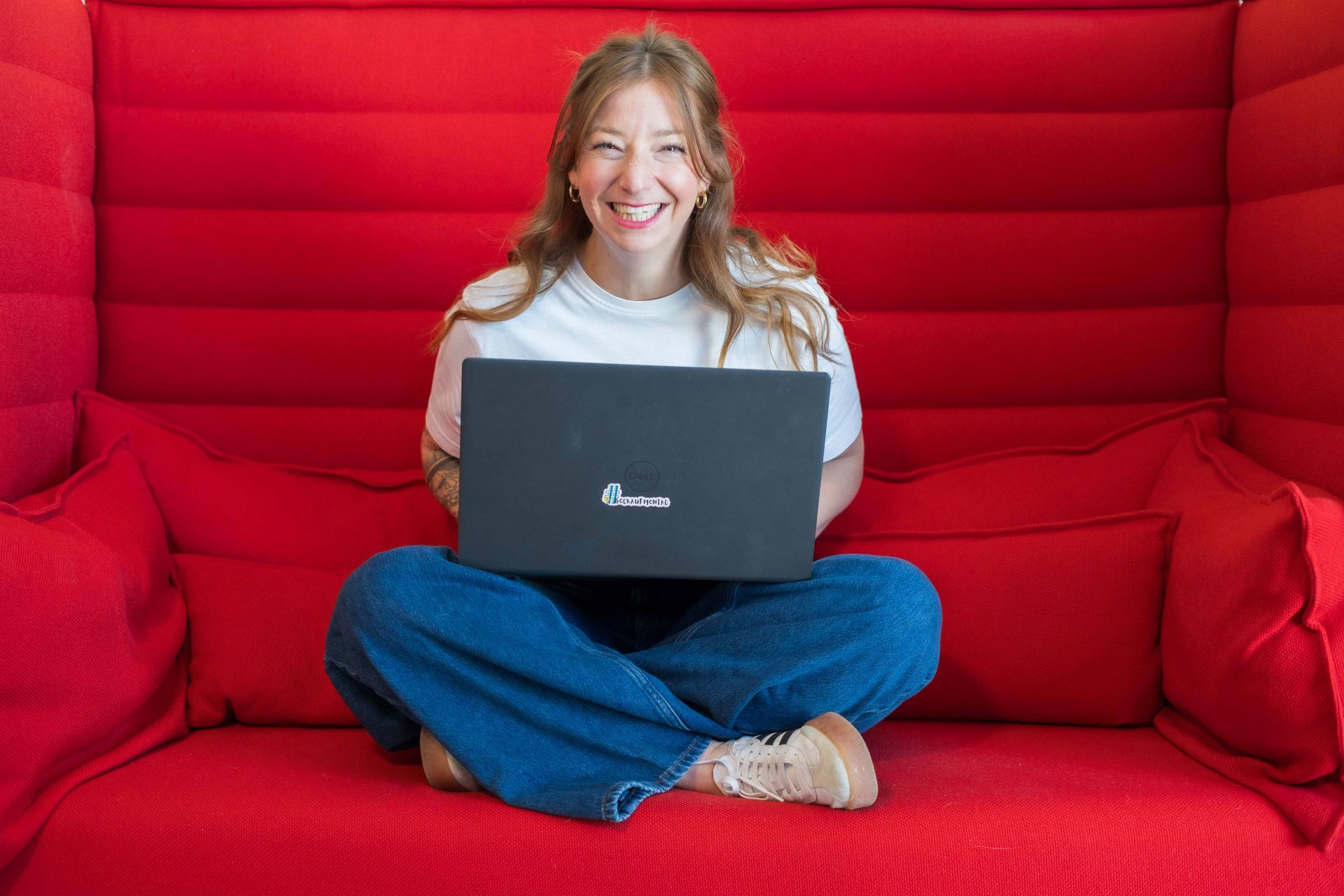 Tanja sitzt lächelnd mit Laptop auf rotem Sofa – Symbolbild für positive Teamkultur, Zusammenarbeit und moderne Führung im Unternehmen.