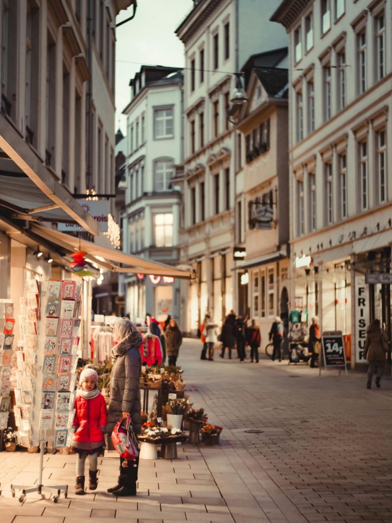 Belebte Einkaufsstraße in einer Innenstadt mit Geschäften, Passanten und weihnachtlich beleuchteten Schaufenstern.