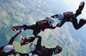 Mehrere Fallschirmspringer fliegen in Formation über einer Landschaft und halten sich in der Luft an den Händen.