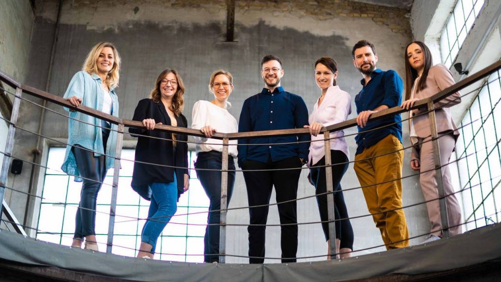 Teambild, auf dem alle gemeinsam auf einer Galerie in einem modernen Industriegebäude stehen und in die Kamera schauen.