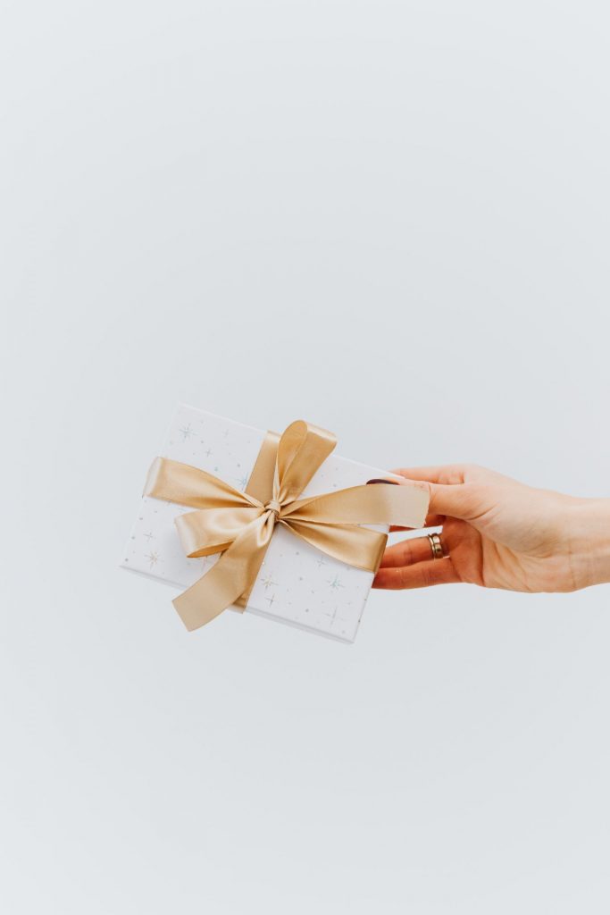 Hand überreicht ein kleines Geschenk mit goldener Schleife vor hellem Hintergrund.