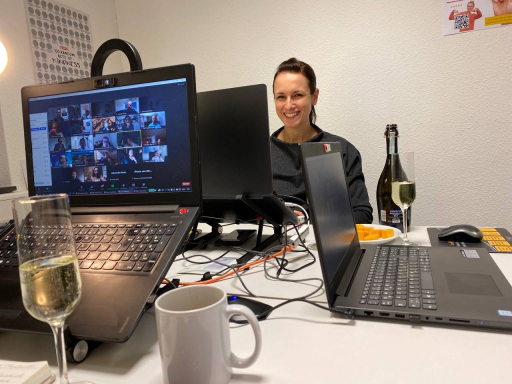 Online-Workshop mit Moderatorin, mehreren Teilnehmenden auf dem Bildschirm und Arbeitsmaterialien am Schreibtisch.