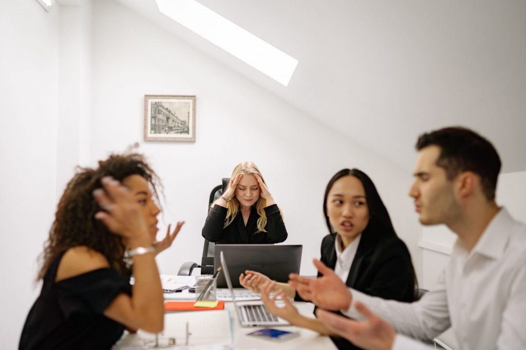 Mehrere Personen diskutieren angespannt in einem Meeting, während eine Kollegin belastet wirkt.