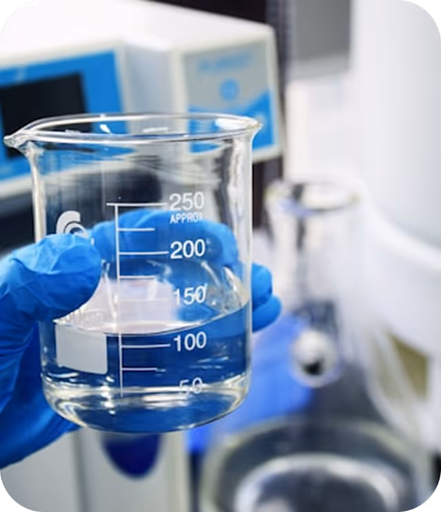 Mano con guante azul sosteniendo un vaso medidor con líquido transparente en un laboratorio.