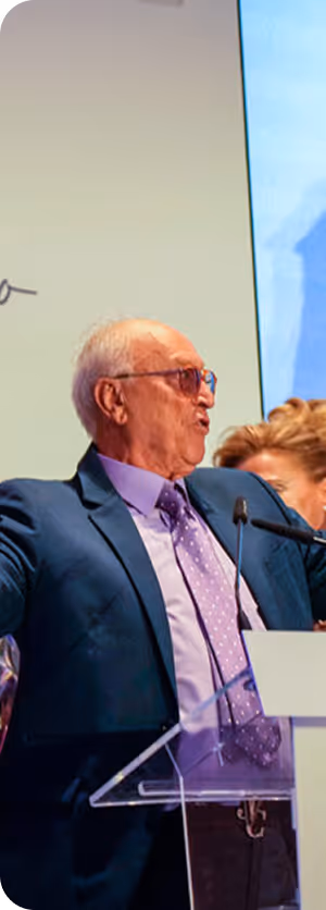 Hombre mayor con gafas y traje hablando en un podio con micrófono en un evento.