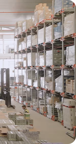 Interior de un almacén con estanterías altas llenas de cajas y un trabajador revisando productos en la parte inferior.