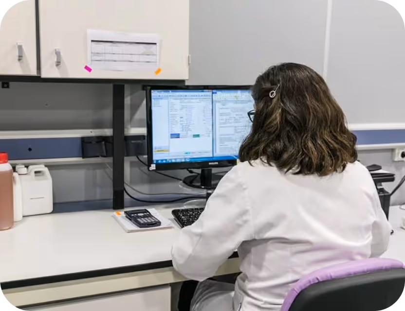Persona con bata blanca sentada frente a un ordenador, trabajando en un entorno de laboratorio o de oficina médica.