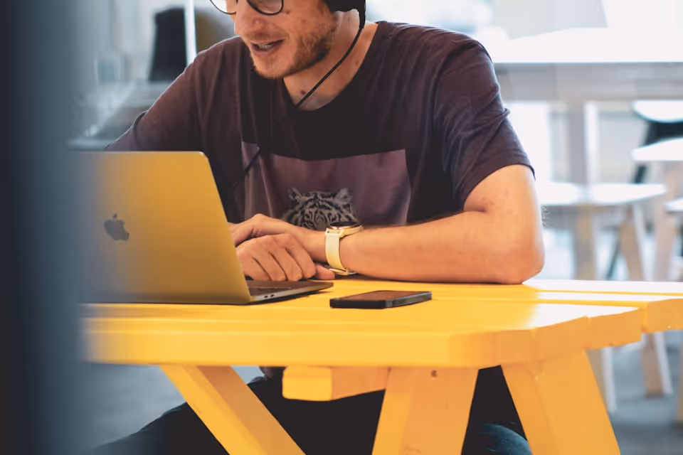 Hombre con gafas y auriculares usando un ordenador portátil Apple en una mesa amarilla con un teléfono móvil.