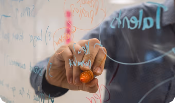 Persona escribiendo con marcador naranja en una pizarra transparente con palabras y diagramas en azul y rojo.