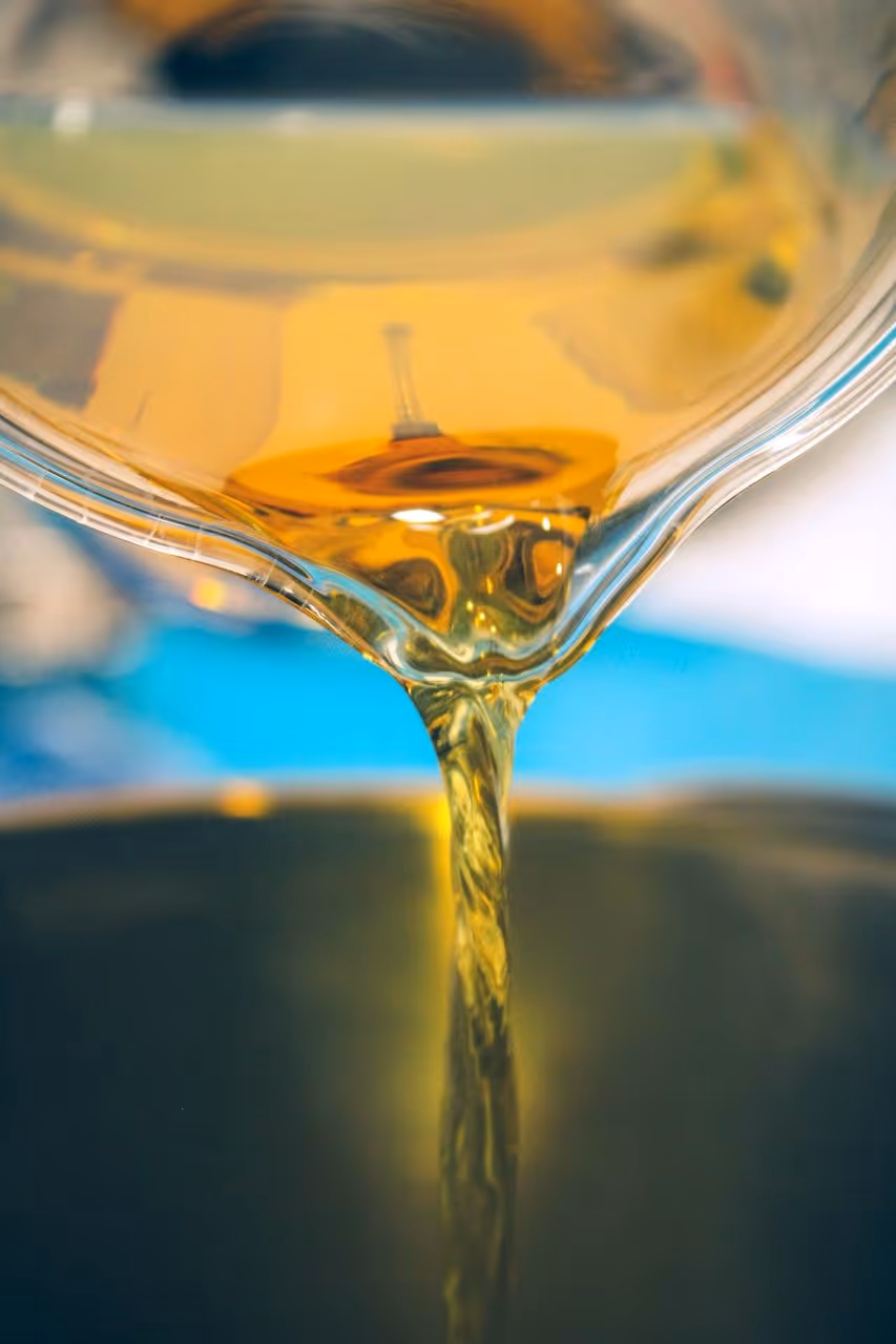 Aceite dorado vertiéndose desde un recipiente transparente hacia una superficie oscura con fondo azul borroso.