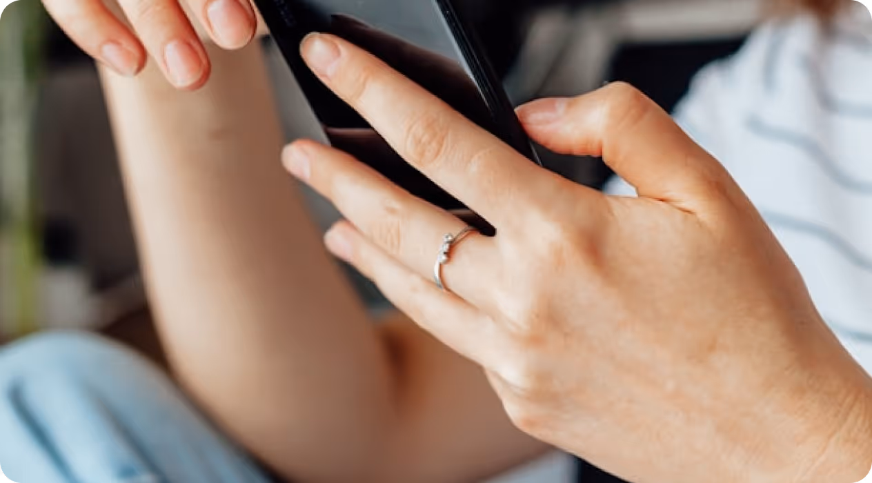 Hands holding a black mobile phone with a silver ring on the ring finger.