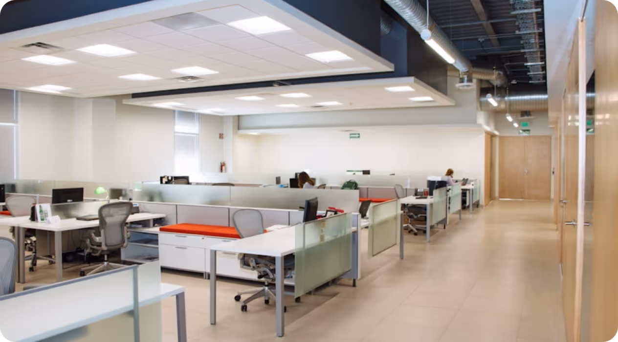 Modern office with white desks, gray ergonomic chairs and glass dividers, with two people working in the background.