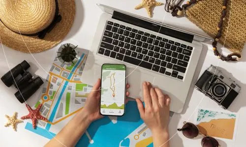 Overhead view of hands holding a smartphone with a hiking route on the screen, surrounded by a laptop, travel map, binoculars, camera, sunglasses, straw hat, starfish, and a woven bag on a white surface.