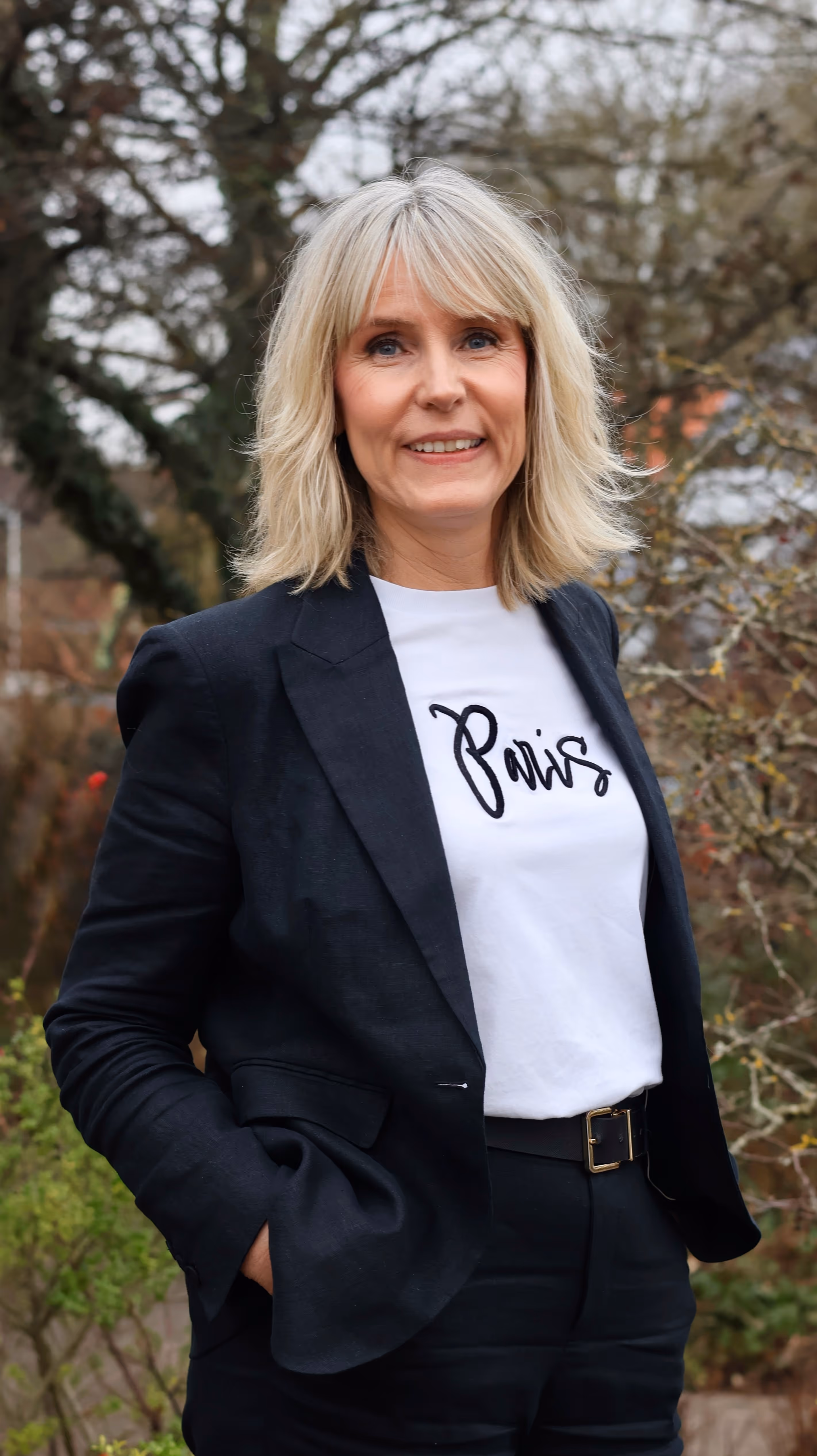 Frau mit blondem Haar trägt schwarzen Blazer und weißes T-Shirt mit der Aufschrift „Paris“ in einem Garten.