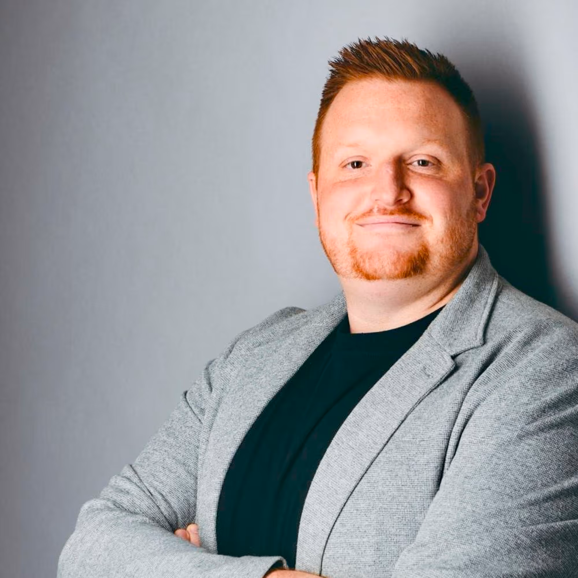 Junge blonde Frau in einem orangefarbenen Blazer, stehend in einem Garten mit Büschen und Bäumen im Hintergrund.