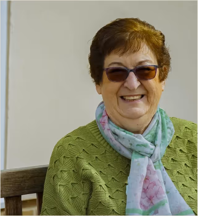Smiling elderly woman wearing sunglasses, a light green knitted sweater, and a floral scarf, sitting on a wooden bench.