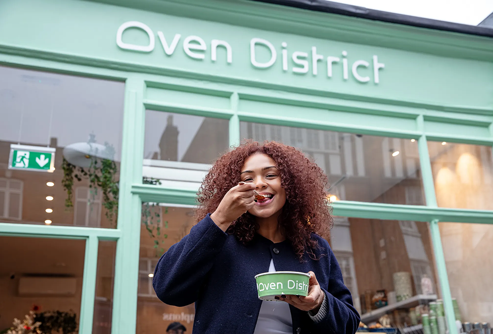 Smiling woman with curly hair eating from a green bowl in front of a mint green storefront reading Oven District.