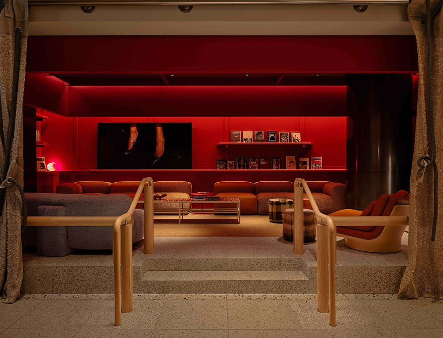 Cozy modern lounge area with red ambient lighting, curved sofas, armchairs, a coffee table, shelves with books, and a large screen on the back wall.