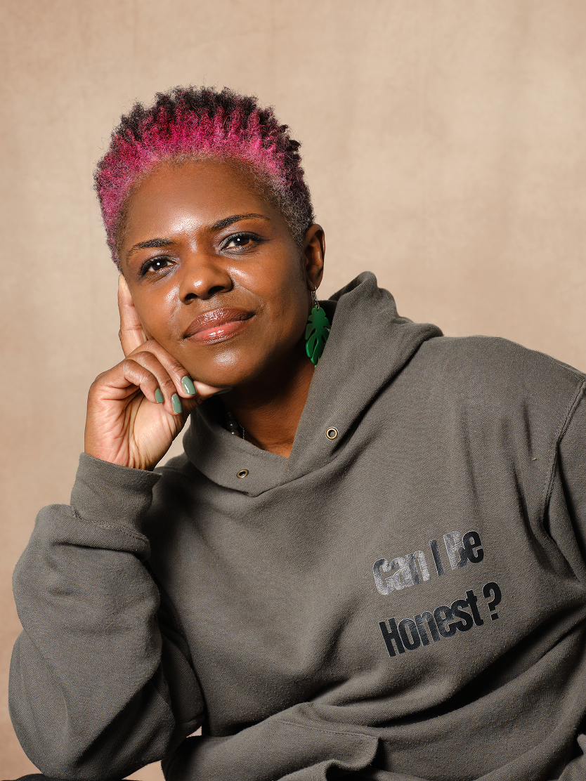 Person with short curly hair dyed pink and black, wearing a gray hoodie with text 'Can I Be Honest?' and green leaf-shaped earrings, resting their head on their hand.