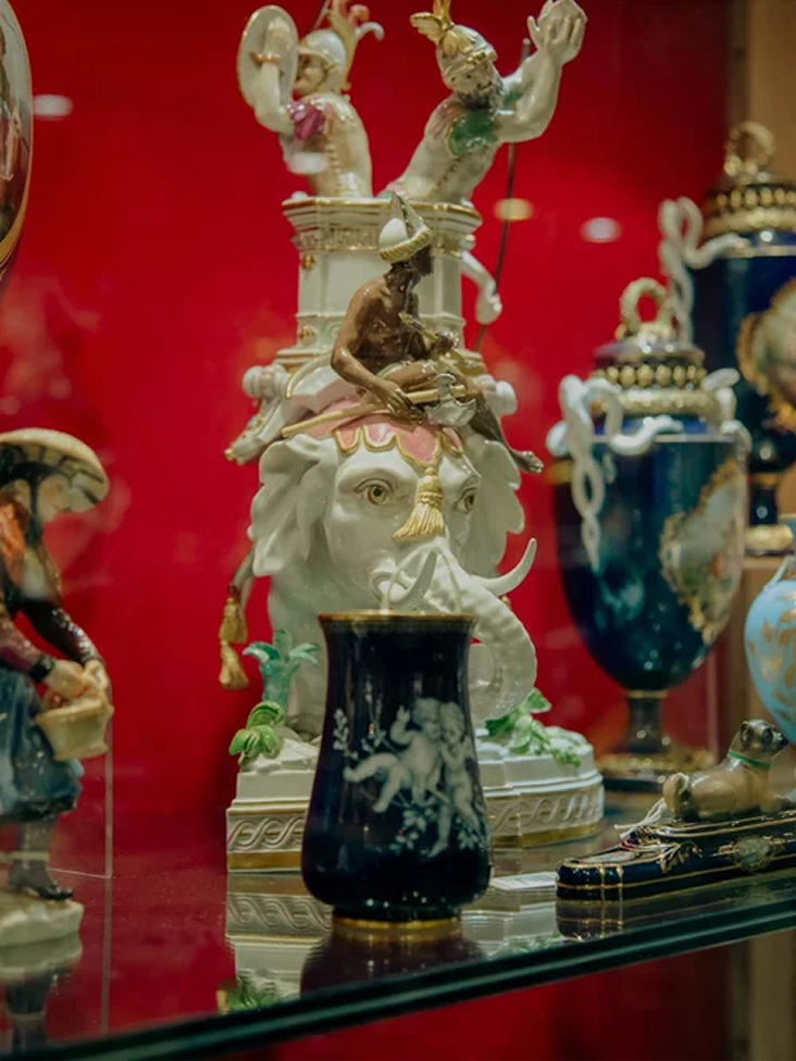 Porcelain figurines and decorative vases displayed on a glass shelf against a red background, including a white elephant with riders and a dark vase with cherub artwork.