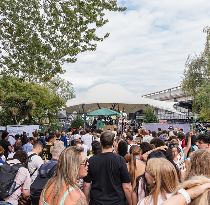 Crowd of people gathered outdoors in front of a white tent stage with trees and a partly cloudy sky in the background.