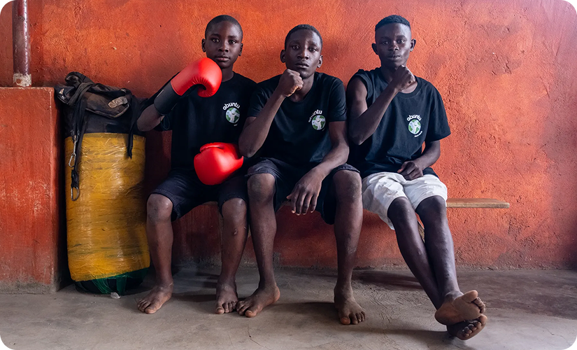 Three boys sitting barefoot on a bench against an orange wall, wearing black t-shirts with a white logo, one boy wearing red boxing gloves.