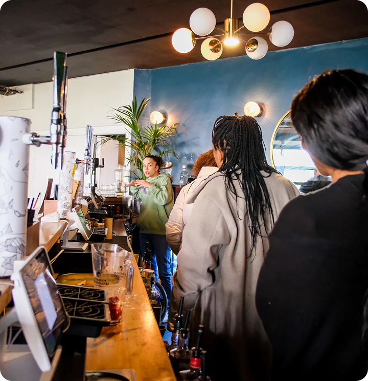 People standing in line at a coffee shop or bar counter with a female barista in green sweater preparing a drink behind the counter.