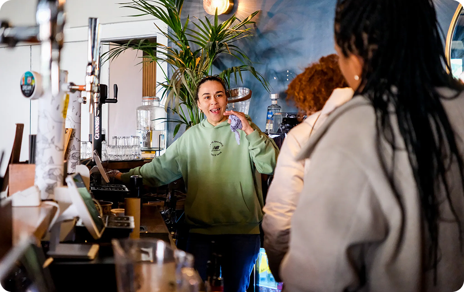 Barista in green sweatshirt talking with two customers at the counter in a cozy café.