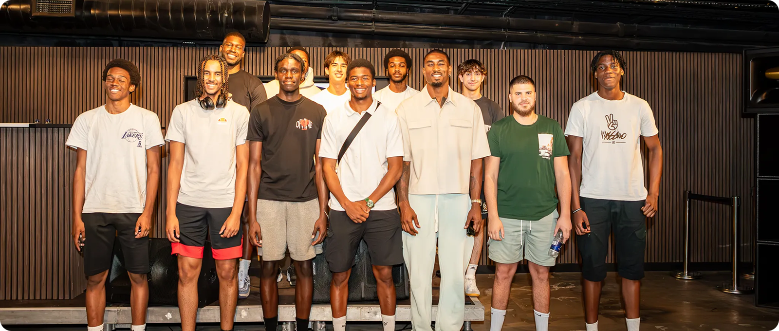 Group of eleven young men standing indoors against a brown slatted wall, smiling toward the camera.