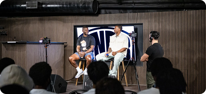 Two men seated on stools having a conversation on a stage in front of an audience, with cameras recording them.