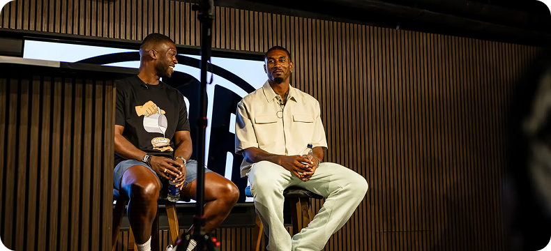 Two men sitting on stools on a stage having a conversation, one wearing a black graphic t-shirt and shorts, the other in a beige shirt and light pants.