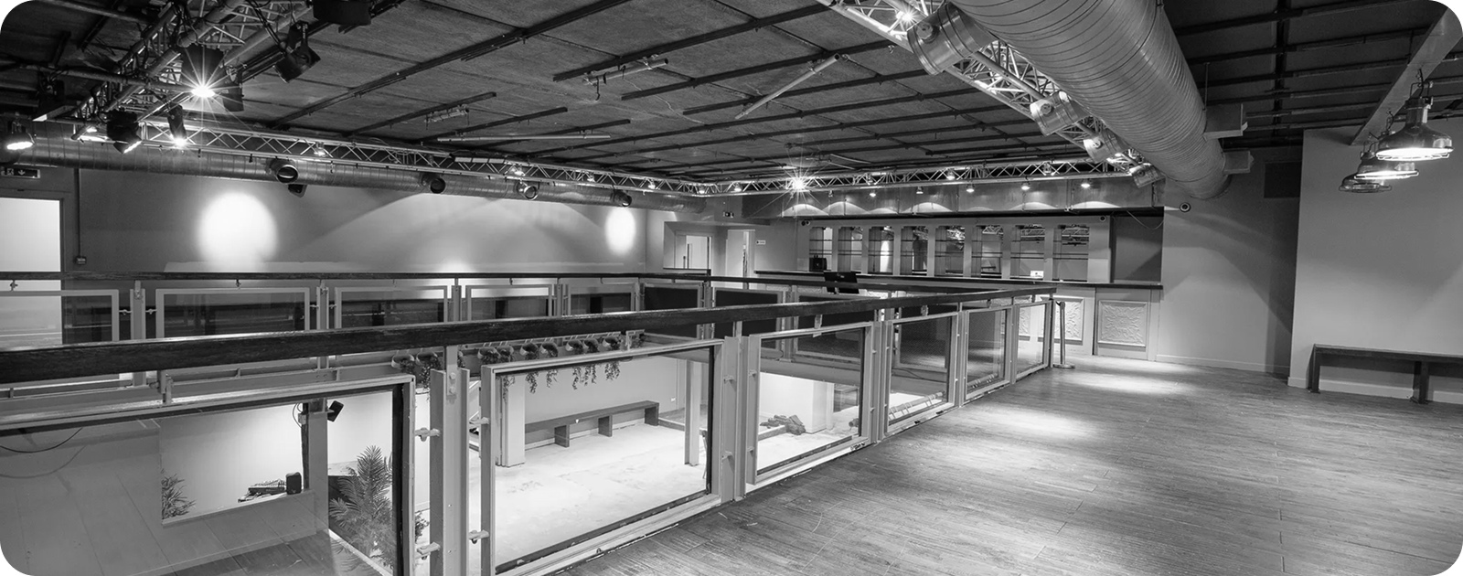 Empty industrial-style interior with mezzanine glass railings, exposed ceiling ductwork, and wooden floor.