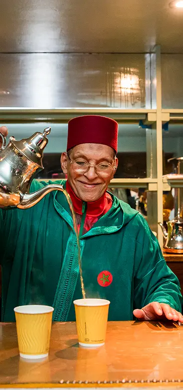Man in red hat and green robe pouring tea from a silver teapot into a paper cup.
